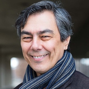 Image of Mario Ortiz-Robles. He has black-grey hair and is wearing a scarf. He is smiling.