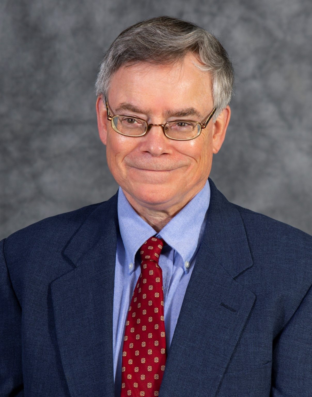 Photograph of Steve Ricke wearing a dark blue suit jacket, light blue dress shirt, and red patterned tie against a gray mottled background. The image appears to be a professional headshot or portrait typically used for business or formal identification purposes.