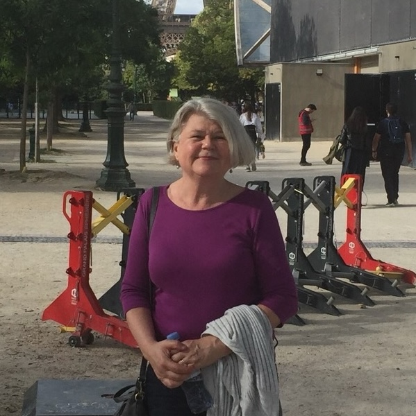 Image of Anne Vila in front of the Eiffel Tower. She is wearing a purple shirt and is holding a jacket. She has short grey hair.