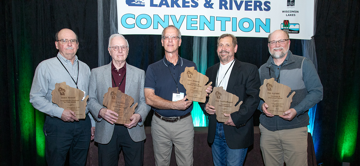 Five smiling men hold wooden plaques in the shape of Wisconsin with a sign hanging on a curtain in the background that reads "LAKES & RIVERS CONVENTION"