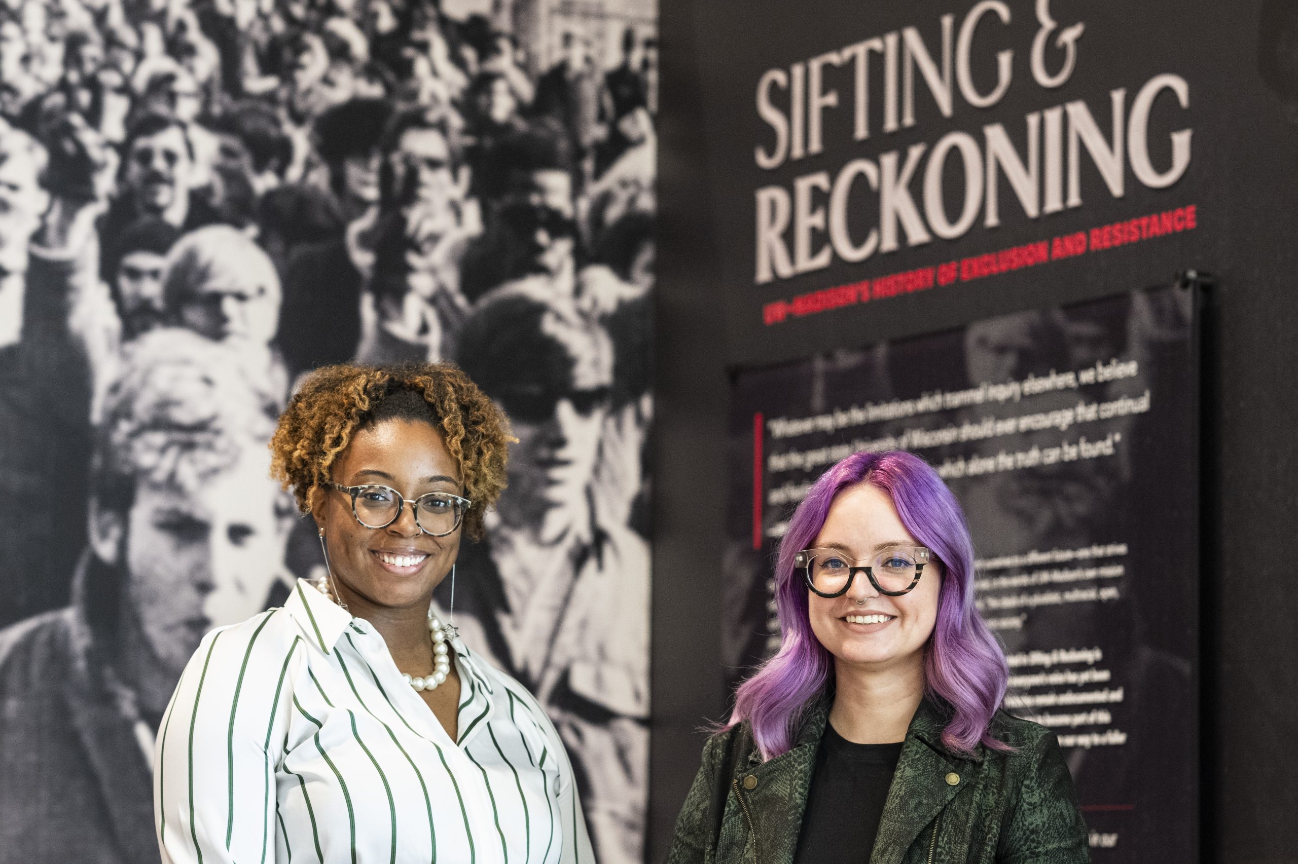 Photo of Taylor L. Bailey and Kacie Lucchini-Butcher standing in front of the large opening panel to the Sifting & Reckoning museum exhibit – a black and white photo of UW–Madison students marching down State Street during the 1969 Black Student Strike.