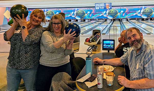 two women hold up bowling balls while a man looks on smiling