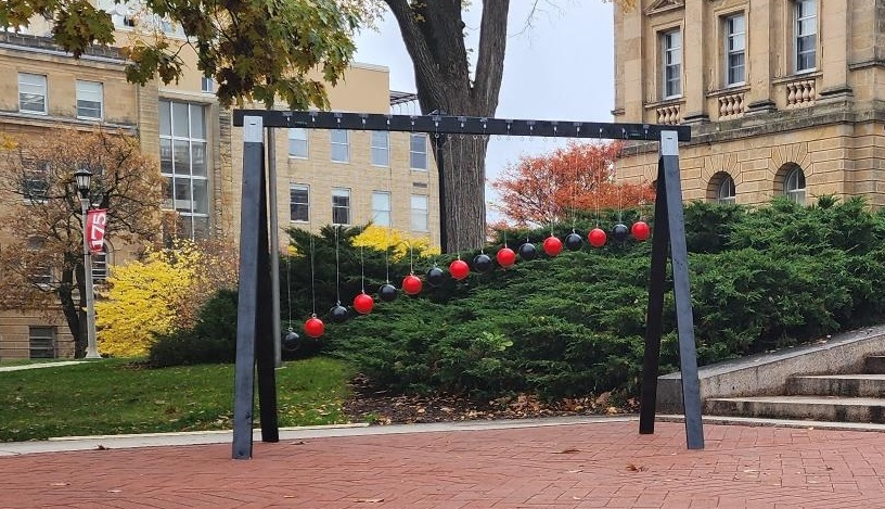 an apparatus that looks a bit like a swingset holds 15 alternating red-black bocce balls from the top cross bar. Each ball is on a string of different length, with the longest at the left and the shortest at the right.