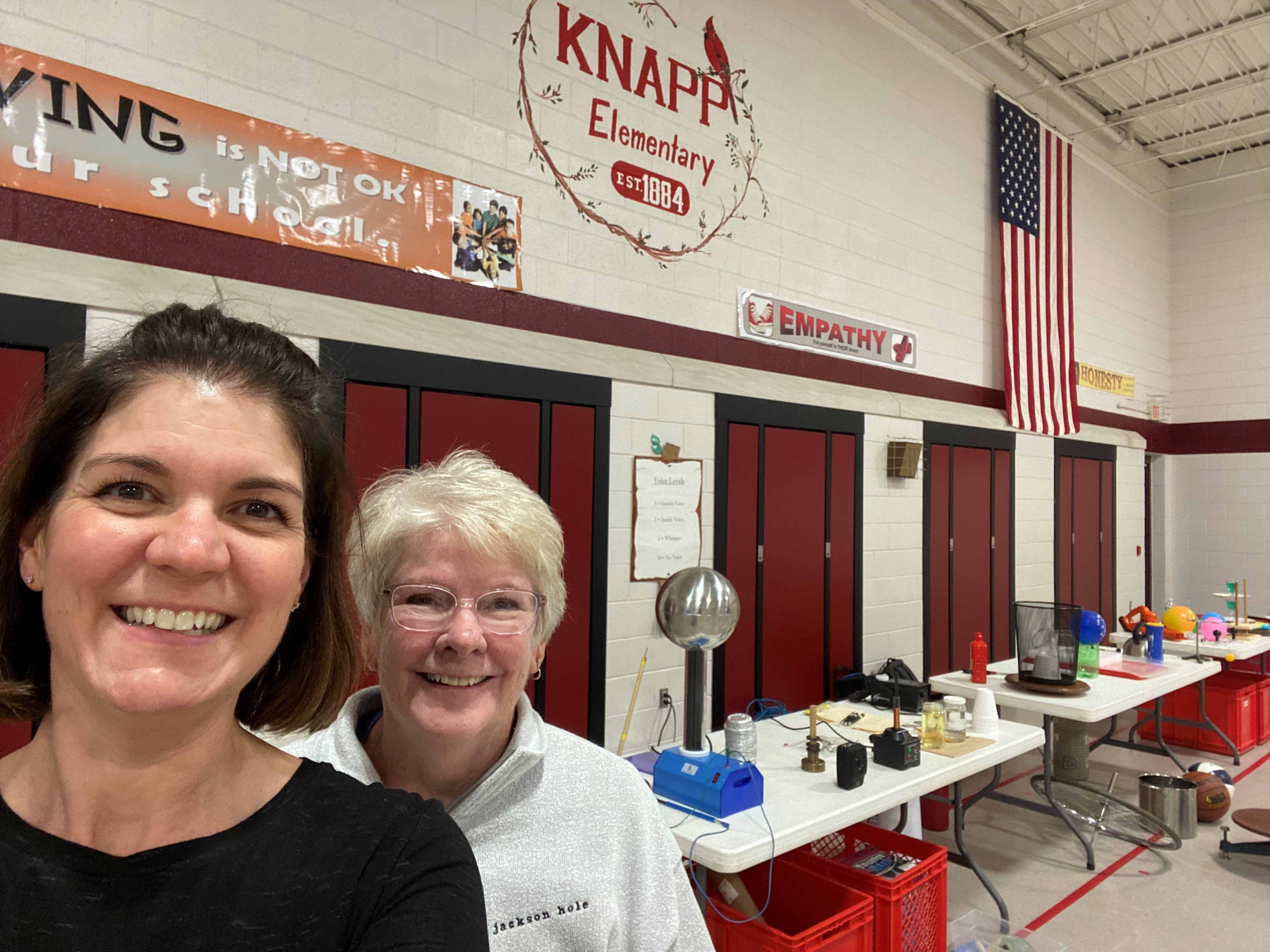 Two women stand in the side of the frame with science demos on tables behind them and a painted "Knapp Elementary" on the wall above them to indicate where they are