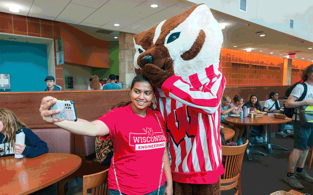 Bucky poses with a student for a selfie in Gordon Dining & Event Center with text overlaid reading "If you want to be a Badger just come along with me"