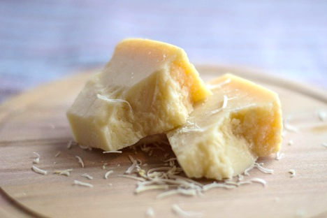 Photograph of two chunks of hard cheese on a wooden cutting board with cheese shavings scattered around. Cheese appears aged with a crumbly texture and pale yellow color, suggesting a rich, savory flavor.