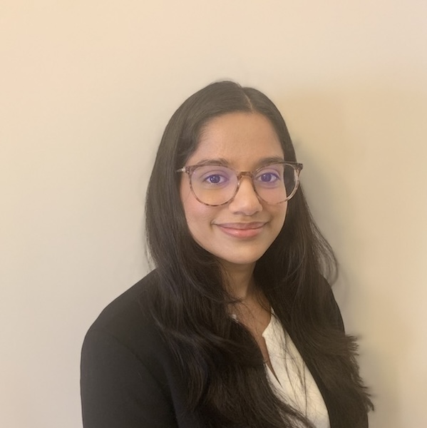 Kuhelika stands in front of a beige background. She has long dark hair and is wearing a black blazer and glasses.