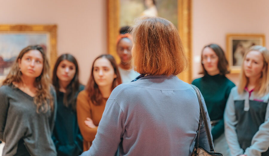Photo of a group of students listening to a Chazen Museum staff member as they stand in front of several paintings in a museum gallery.