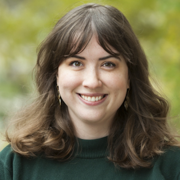 Allyson stands in front of blurred greenery. She has medium length brown hair and is wearing a dark green sweater.