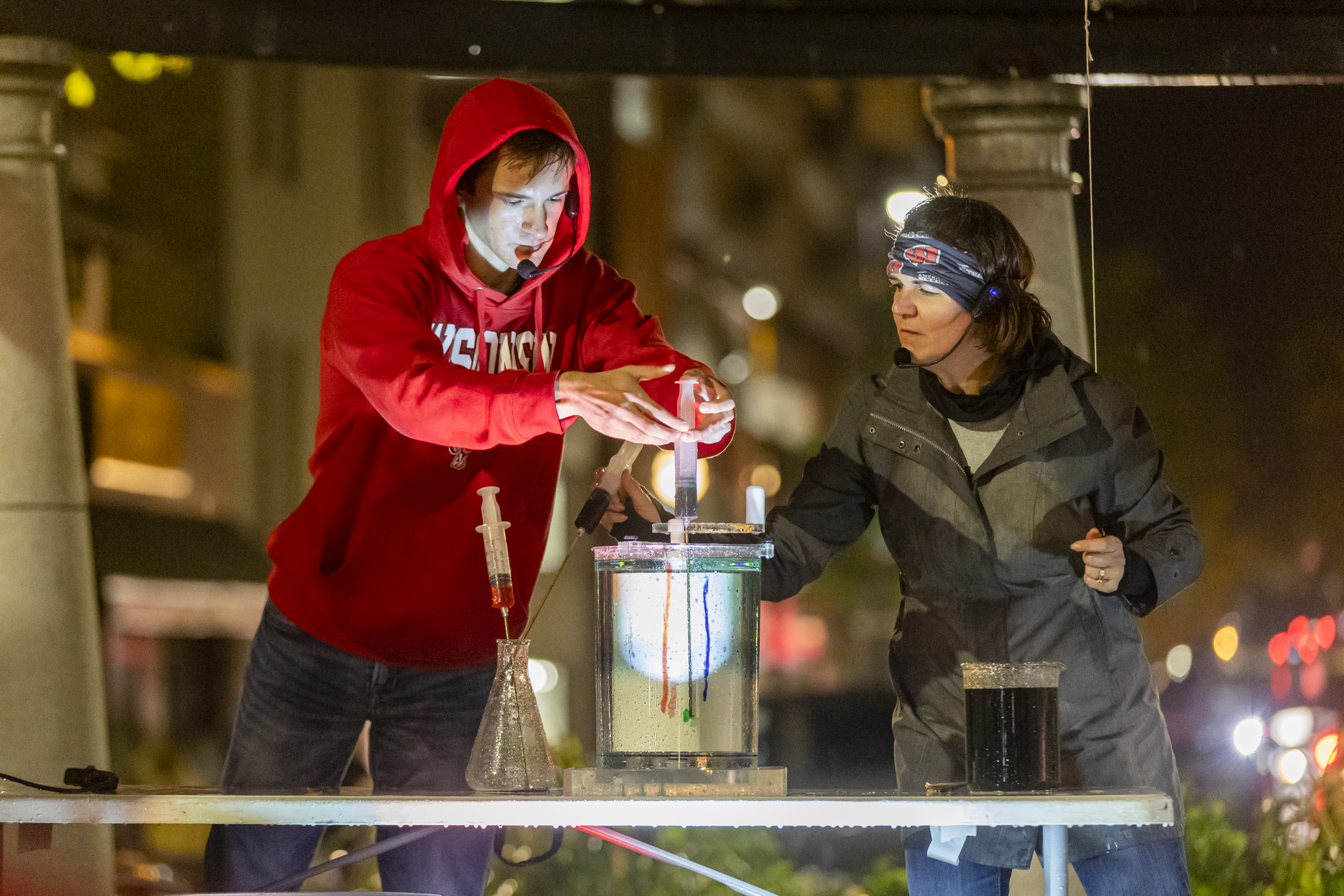 a man in a red Wisconsin sweatshirt uses a syringe to inject colored liquid into a large beaker. A woman, bundled up from the rain and cold, looks on