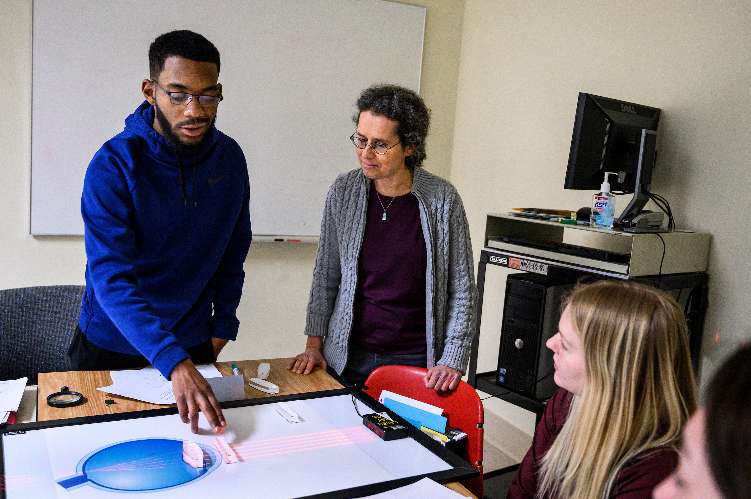 Photo of Susan Nossal, director and founder of the Physics Learning Center, standing and listening to a student discussing an optical calculation he is working through on a large whiteboard on a table as other students listen. 