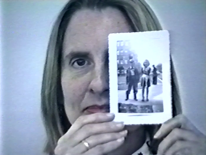 A woman holds up a photo in front of half of her face. The photo shows her as a child next to another child.