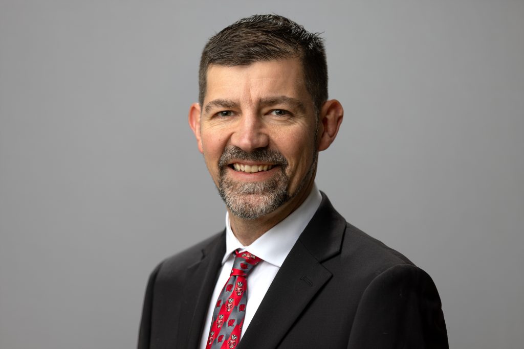 Photo of Jeff Sindelar in red tie, white shirt and black jacket on a gray background