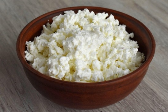Photograph of a wooden bowl filled with white, crumbly cottage cheese placed on a light wooden surface. The image highlights texture and freshness of cottage cheese as a dairy product.