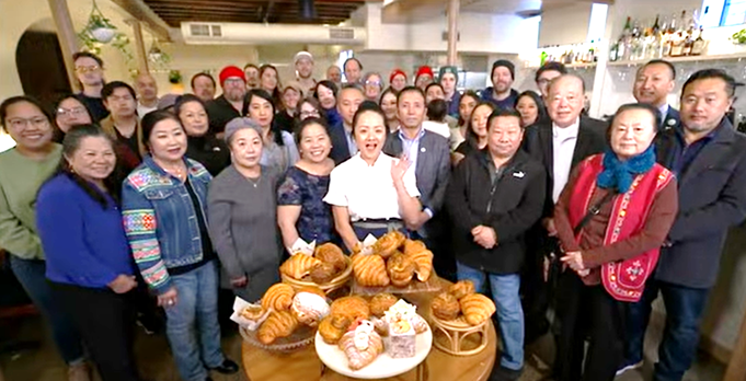 Photograph of a large group of people gathered around a table filled with various types of bread and pastries inside Diane's Place restaurant. At center is Diane Moua, surrounded by family and friends, including Houa Moua off to the right.