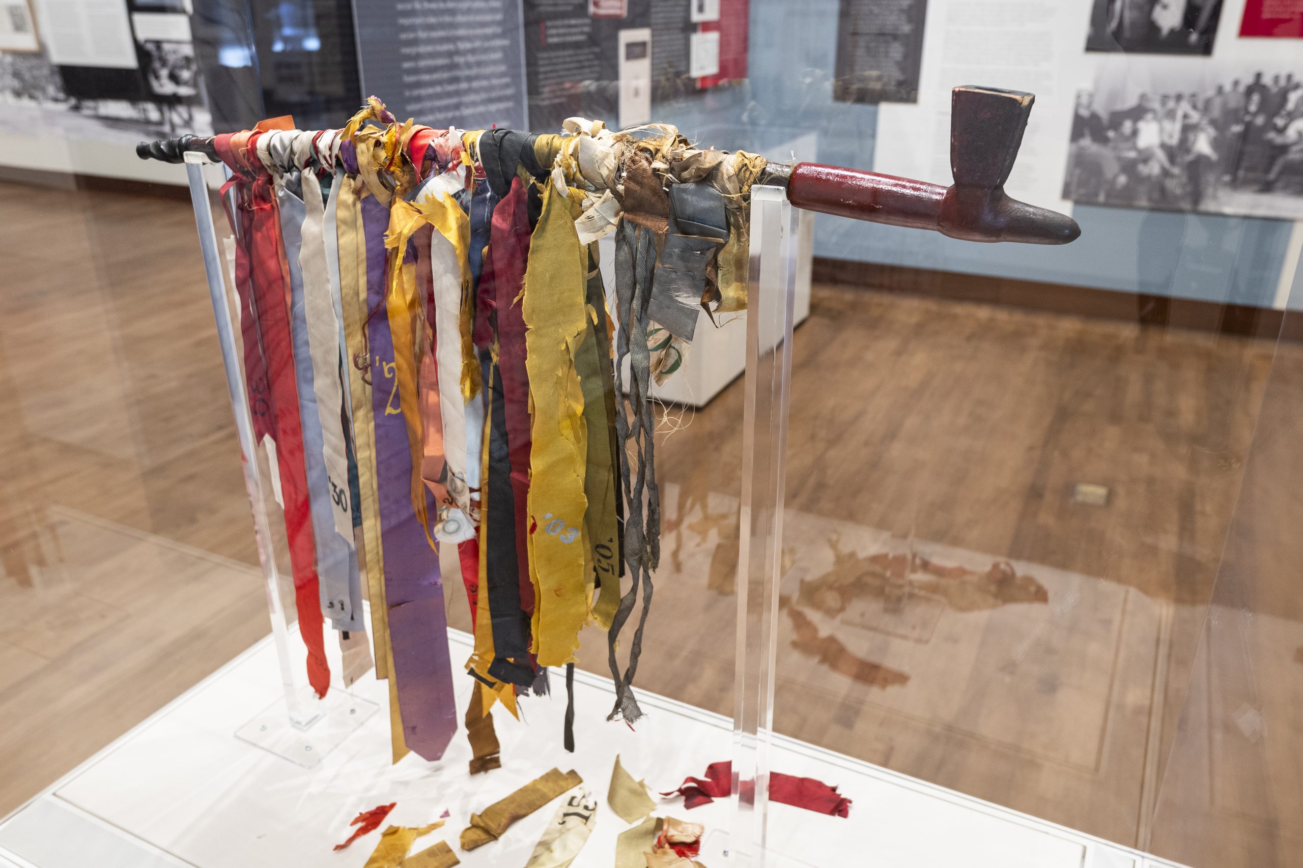 Photo of the pipe of peace, a ceremonial object used by white students in a popular mock Native ceremony in the early 20th century. It is a carved wooden pipe about two feet long around which have been tied ribbons of different colors representing different class years.