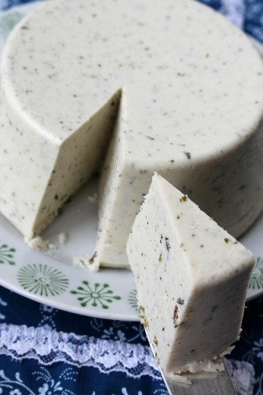 Photograph of a round, white, speckled cashew cheese with a wedge cut out and lifted on a spatula. The item rests on a white plate with green floral patterns, placed on a blue lace tablecloth, highlighting texture and consistency.