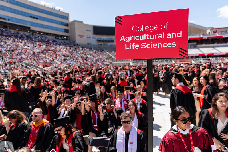 A large group of graduates at Camp Randall stadium in Madison, Wis. with a red sign in the foreground with the message "College of Agricultural and Life Sciences"